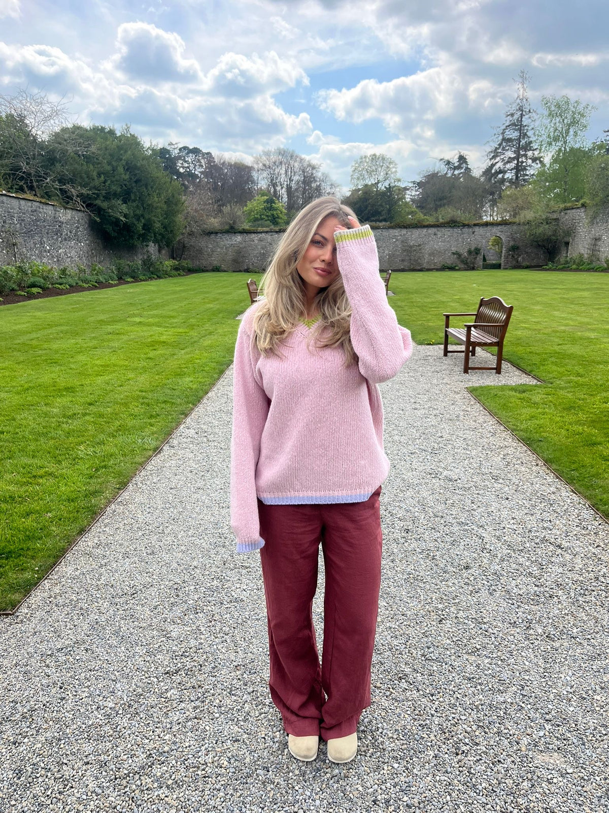 Gemma Cowen standing on a gravel path in a garden wearing a pink V-neck wool aran jumper with green trim on the sleeves, paired with maroon trousers.
