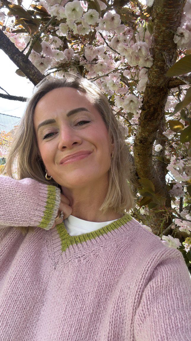 Woman wearing a pink sweater with green trim standing in front of cherry blossom trees.